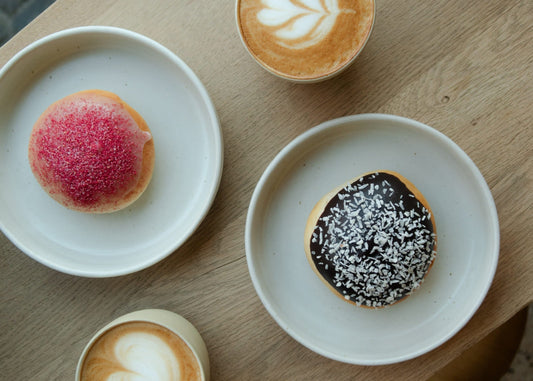 Flatlay-billede på et lyst træbord med to forskellige fastelavnsboller og to kopper cappuccino med latte art.