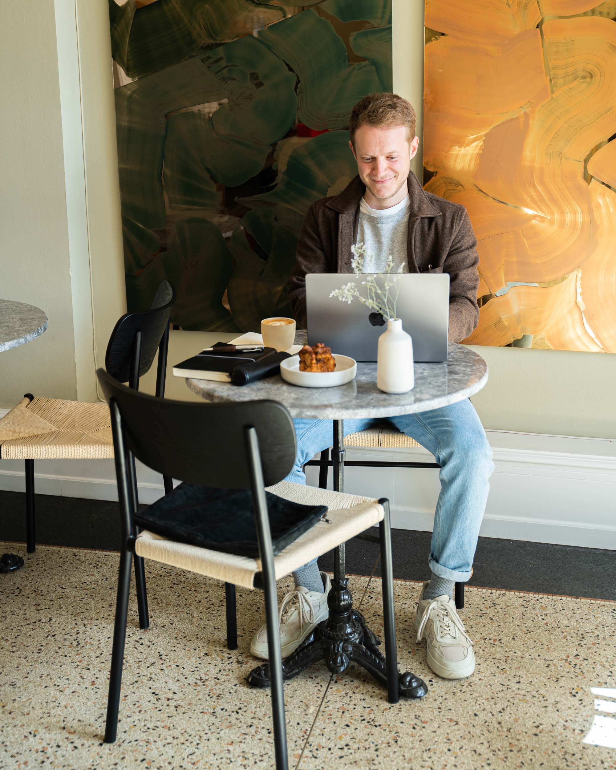En mand sidder ved et bord med en bærbar computer foran sig på KALA og nyder en kop kaffe